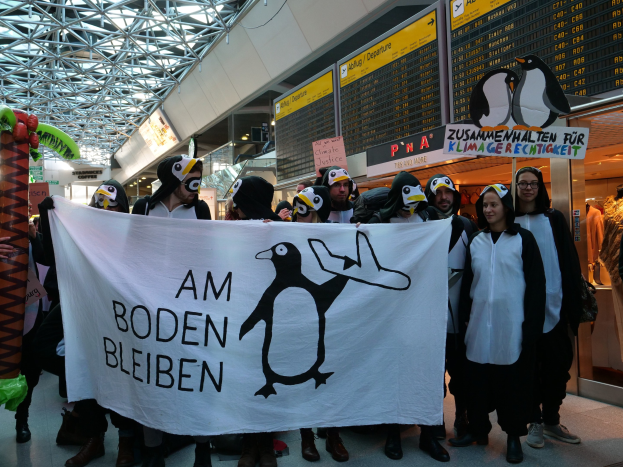 Gruppe von Menschen in Pinguinkostümen mit einem Banner vor einer Wand mit Pinguin-Plakaten, Schaufensterpuppen mit Kleidern und anderen Gegenständen im Hintergrund.